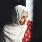 A thoughtful portrait of a woman in a hijab sitting indoors, bathed in natural light.